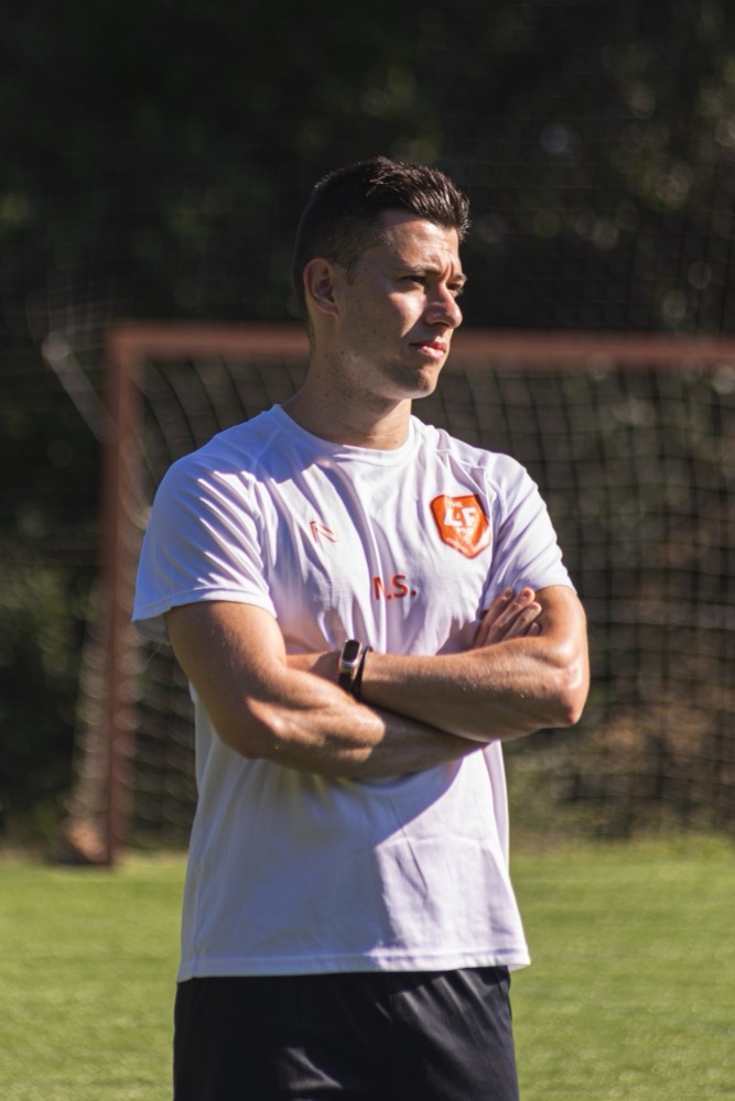 Mario Sabou, con la camiseta blanca de THEL4FPRO, observando un entrenamiento en el campo de la academia